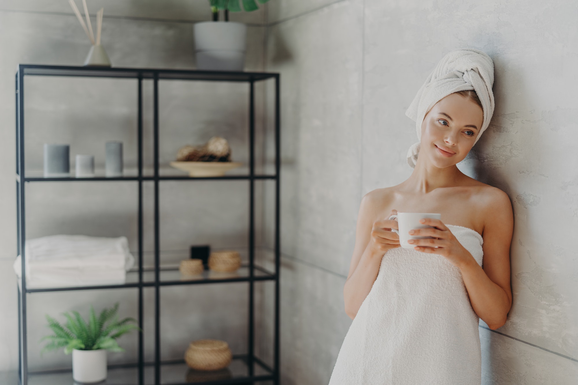 Calm relaxed woman poses in towel against bathroom interior, wrapped in bath towel